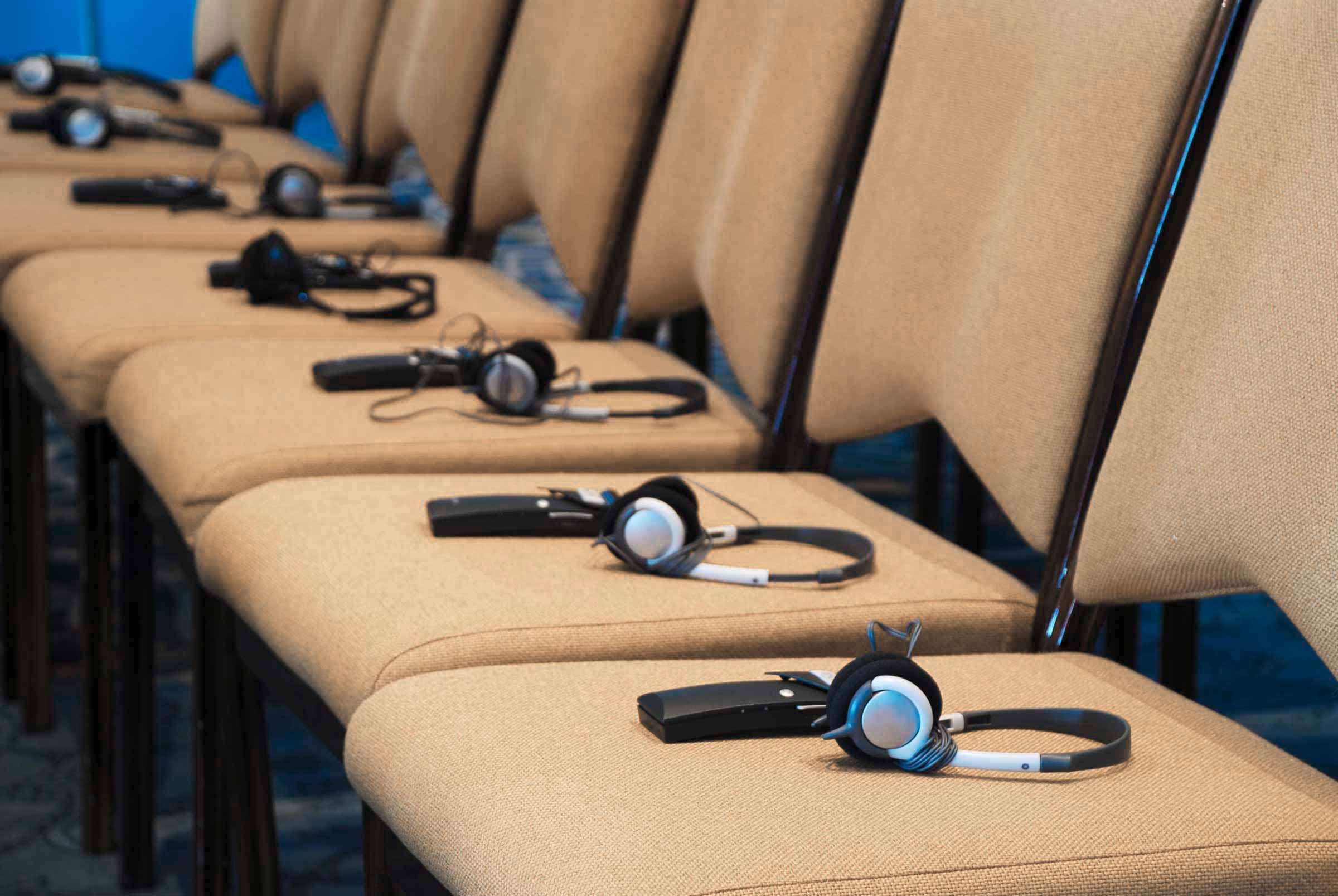 Nahaufnahme einer Stuhlreihe bei einer Konferenz. Auf jeder Sitzfläche liegt ein Empfangsgerät und ein Kopfhörer für den Empfang der Simultandverdolmetschung.