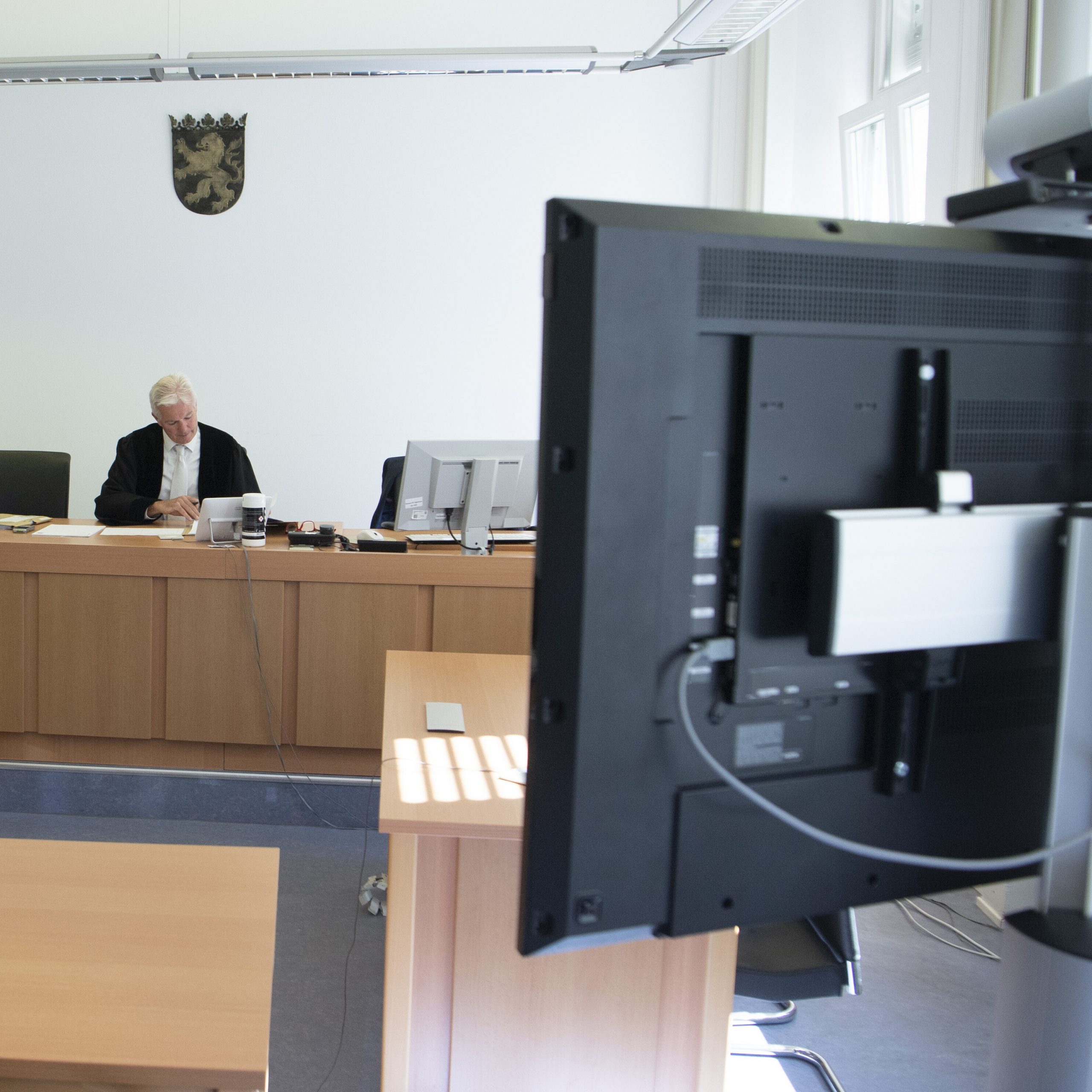Im Gerichtssaal. Links der Richtertisch, an dem ein Richter sitzt. Rechts im Vordergrund der Bildschirm eines Videokonferenzsystems von hinten. Die Kamera auf dem Bildschirm ist auf de Richter gerichtet.
