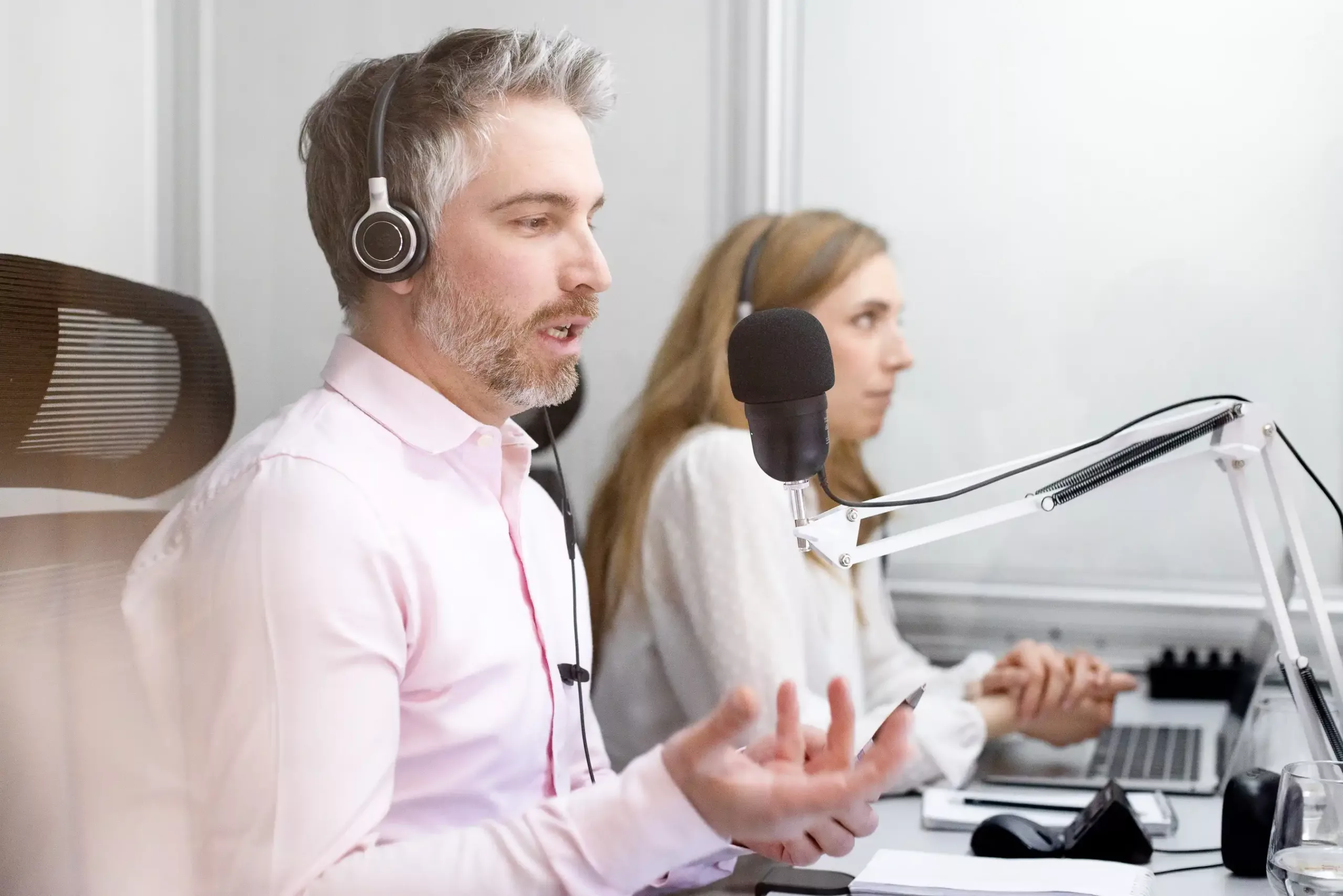 Im Vordergrund eine Simultanübersetzer bei der Arbeit in der Dolmetschkabine. Er hat Kopfhörer auf und spricht in ein Mikrofon, das auf einem Mikrofonarm steckt. Neben ihm in Hintergrund die Teamkollegin vor ihrem Laptop.