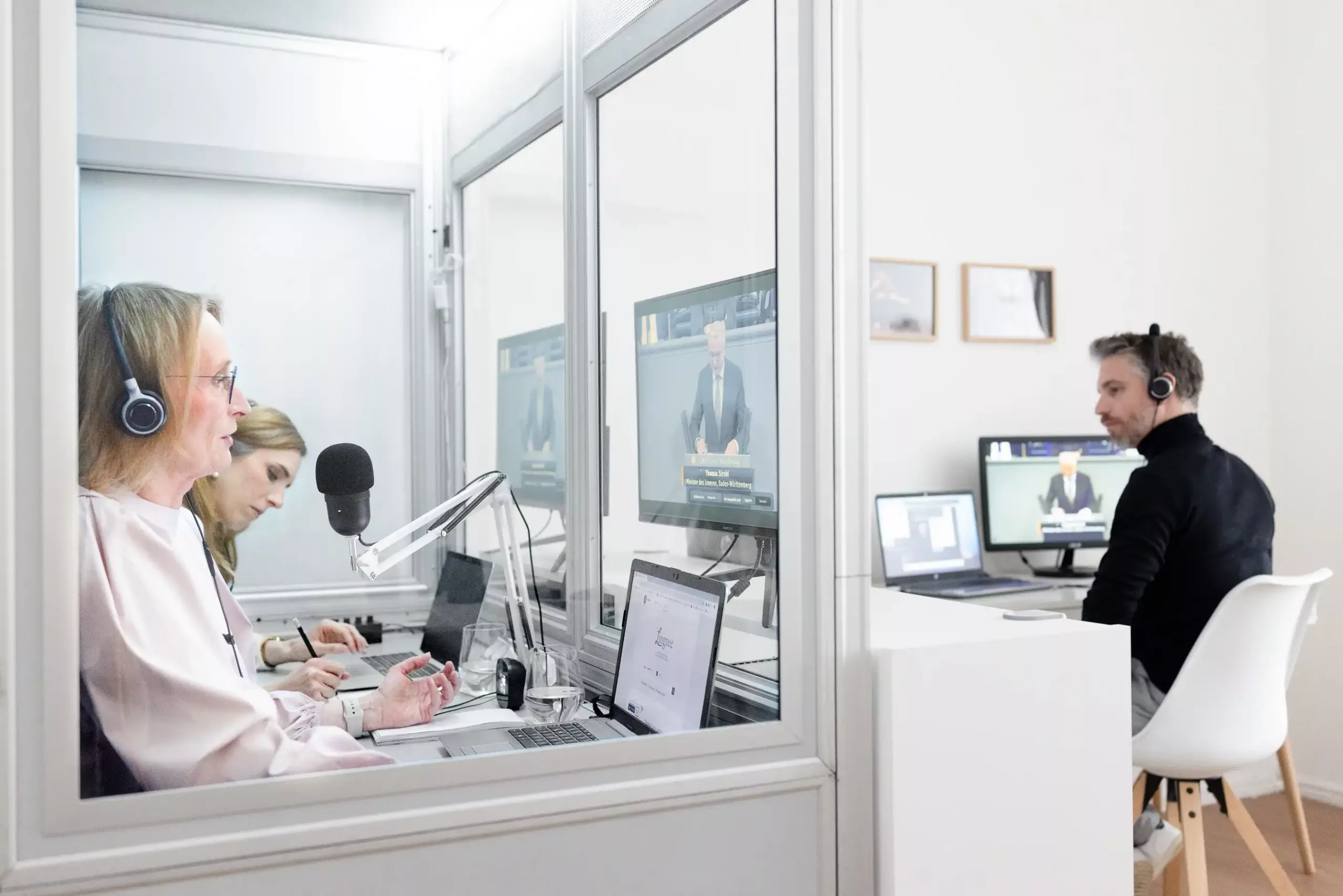 Links im Bild zwei Dolmetscher:innen in einer mobilen Dolmetschkabine bei der Arbeit. Sie kaben Kopfhörer auf und sprechen in ein schwarzes Mikrofon, das auf einem weißen Mikrofonarm steckt. Sie schauen auf Bildschirme, die vor der Kabine stehen. Darauf ist ein Redner im deutschen Bundestag zu sehen. Rechts im Bild an einem Schreibtisch der Techniker des Dolmetschstudios. Er sitzt vor einem Bildschirm und einem Laptop und hat einen Kopfhörer auf. Aufmerksam überwacht er die Technik.